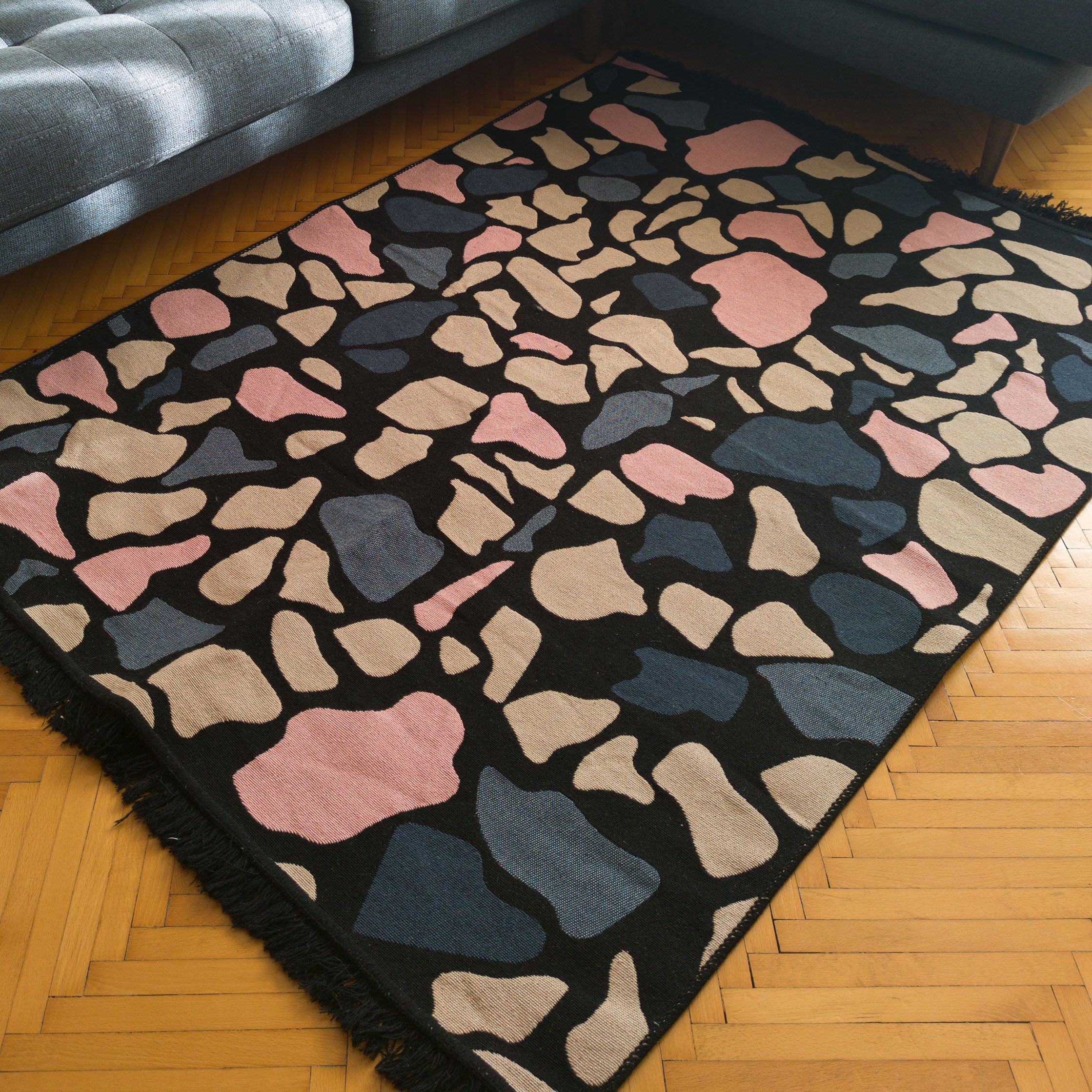 Abstract patterned rug with pink, beige, and blue shapes on black background, styled in living room with wooden floor